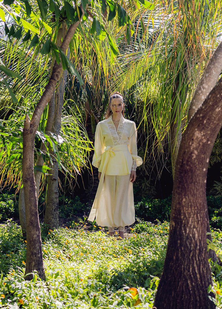 Look completo em amarelo manteiga, com faixa na cintura e calça de barra bufante, fotografado em jardim tropical.