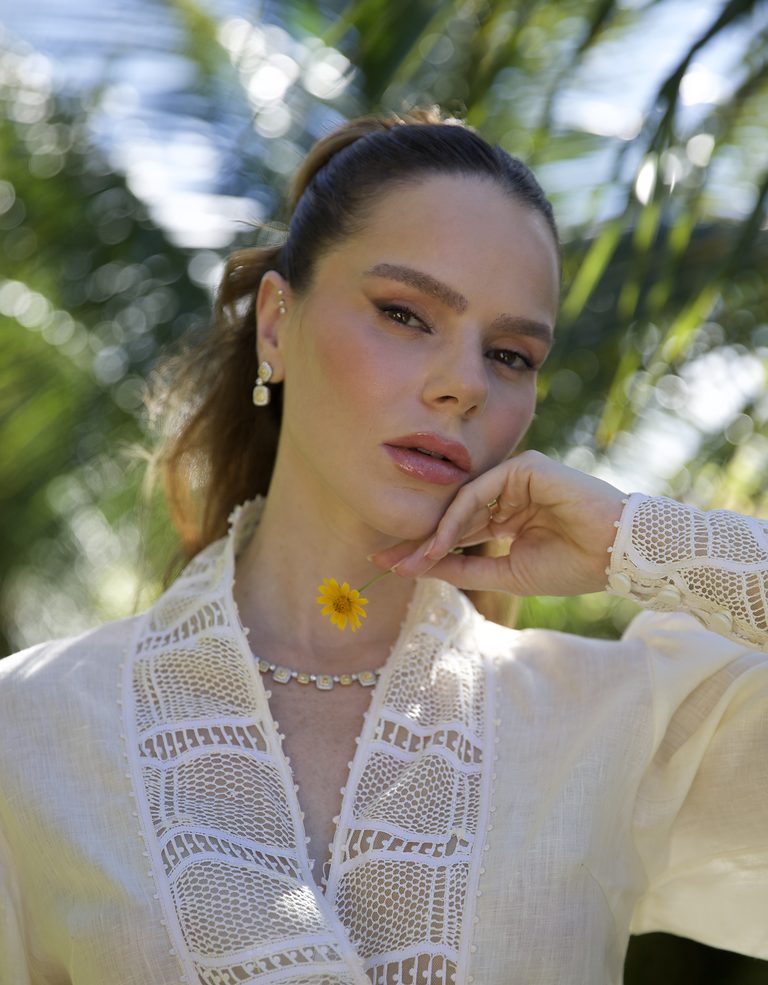 Retrato de modelo com blusa de linho e renda renascença, segurando uma flor amarela.