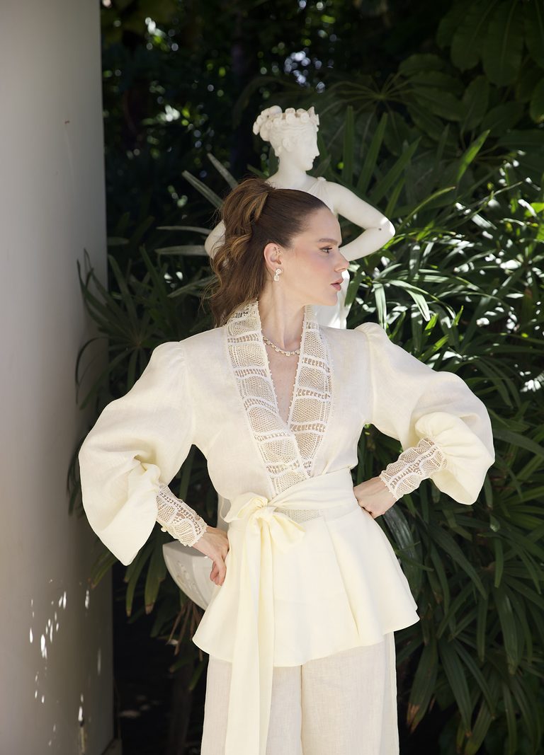 Modelo veste blusa de linho manteiga com gola e punhos em renda renascença, em jardim com escultura de mármore.